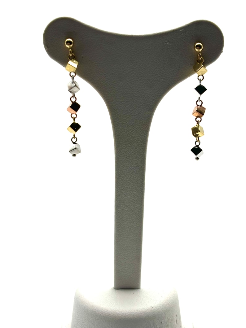 Op de afbeelding: Een paar gouden, zilveren en ros&eacute;gouden oorhangers met een ketting van kleine vierkante kralen.