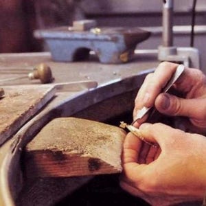 Pode incluir: Um close-up das m&atilde;os de uma pessoa trabalhando em uma pe&ccedil;a de joia com uma pequena ferramenta. A joia est&aacute; em uma superf&iacute;cie de madeira e a pessoa est&aacute; vestindo uma camisa marrom.