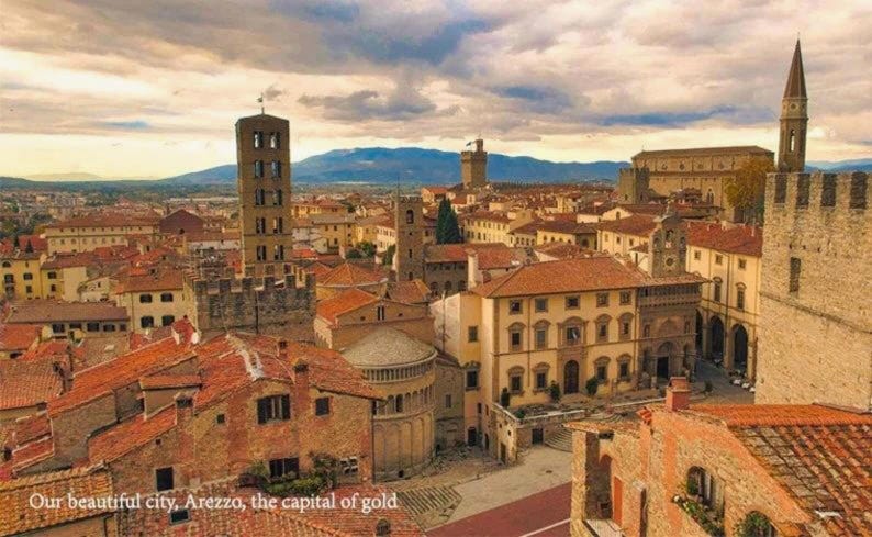 Pode incluir: Uma vista a&eacute;rea de uma cidade italiana hist&oacute;rica com edif&iacute;cios de cor terracota, uma torre alta e uma grande muralha de pedra. A cidade &eacute; cercada por colinas onduladas e um c&eacute;u nublado.