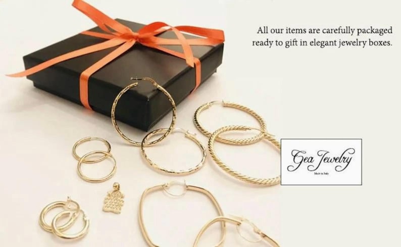 Pode incluir: Um conjunto de brincos de argola dourados em v&aacute;rios tamanhos e estilos, incluindo um par de brincos quadrados. Os brincos s&atilde;o exibidos em uma caixa de presente preta com uma fita vermelha. O texto "Gea Jewelry" &eacute; vis&iacute;vel na imagem.