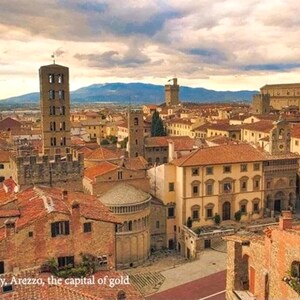 Op de afbeelding: Een luchtfoto van een historische Italiaanse stad met terracotta daken, een hoge klokkentoren en een groot stenen gebouw op de achtergrond. De stad wordt omringd door heuvels en bergen.