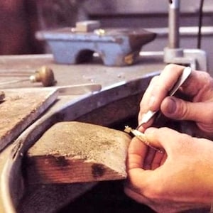 Op de afbeelding: Een close-up van de handen van een juwelier die een pincet gebruikt om aan een stuk gouden sieraden te werken op een houten werkbank.
