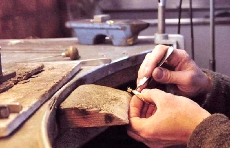 Op de afbeelding: Een close-up van de handen van een juwelier die een pincet gebruikt om aan een stuk gouden sieraden te werken op een houten werkbank.