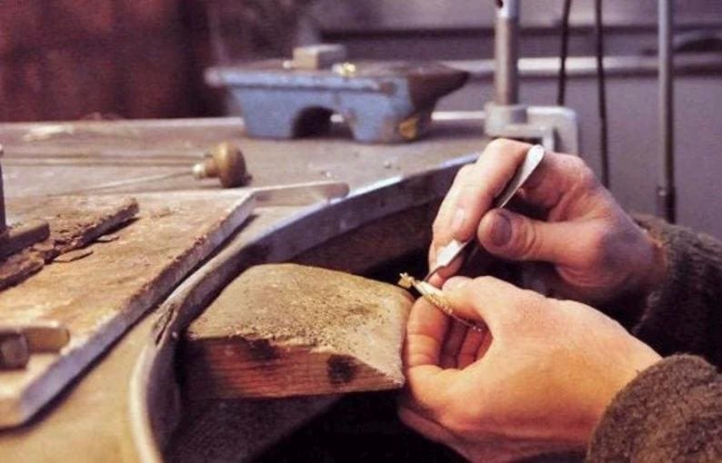 May include: A close-up of a person's hands using tweezers to work on a piece of jewelry on a wooden surface. The jewelry is gold and has a delicate design.