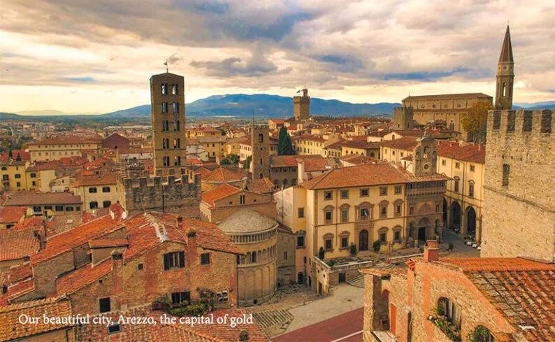 May include: An aerial view of a historic city with terracotta-colored buildings, a tall tower, and a large stone wall. The city is surrounded by rolling hills and a cloudy sky.