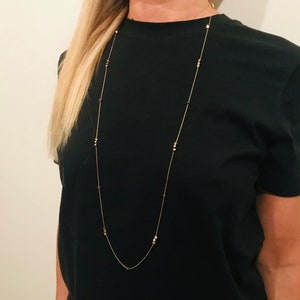 Op de afbeelding: Een lange gouden ketting met kleine ronde kralen. De ketting wordt gedragen door een persoon met lang blond haar en een zwart T-shirt.