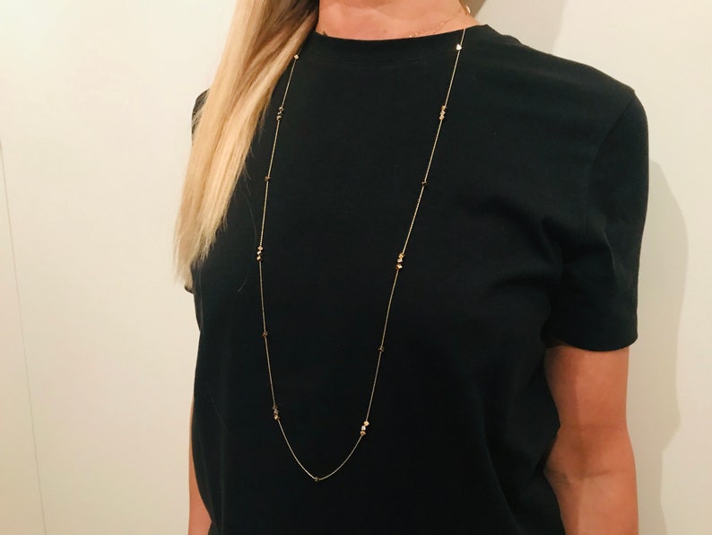 Op de afbeelding: Een lange gouden ketting met kleine ronde kralen. De ketting wordt gedragen door een persoon met lang blond haar en een zwart T-shirt.