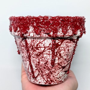 May include: A white ceramic planter with red paint splatters and drips. The rim of the planter is covered in red paint and small red crystals.