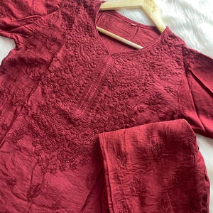 Puede incluir: Un conjunto de dos piezas de color granate, que incluye una parte superior de manga larga y pantalones a juego. La parte superior presenta intrincados detalles bordados alrededor del escote y el corpiño. Los pantalones también tienen detalles bordados. El conjunto está colgado en una percha de madera.