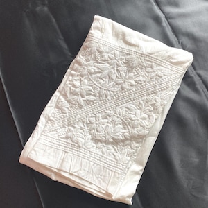 Peut inclure: Un tissu blanc plié avec une broderie florale complexe. Le motif détaillé recouvre une partie du tissu, avec un bord plissé visible. Le tissu repose sur un fond gris foncé, possiblement satiné.