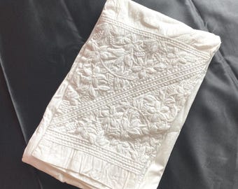 May include: A folded white fabric with intricate floral embroidery. The detailed design covers a portion of the fabric, with a pleated edge visible. The fabric rests on a dark gray, possibly satin, background.