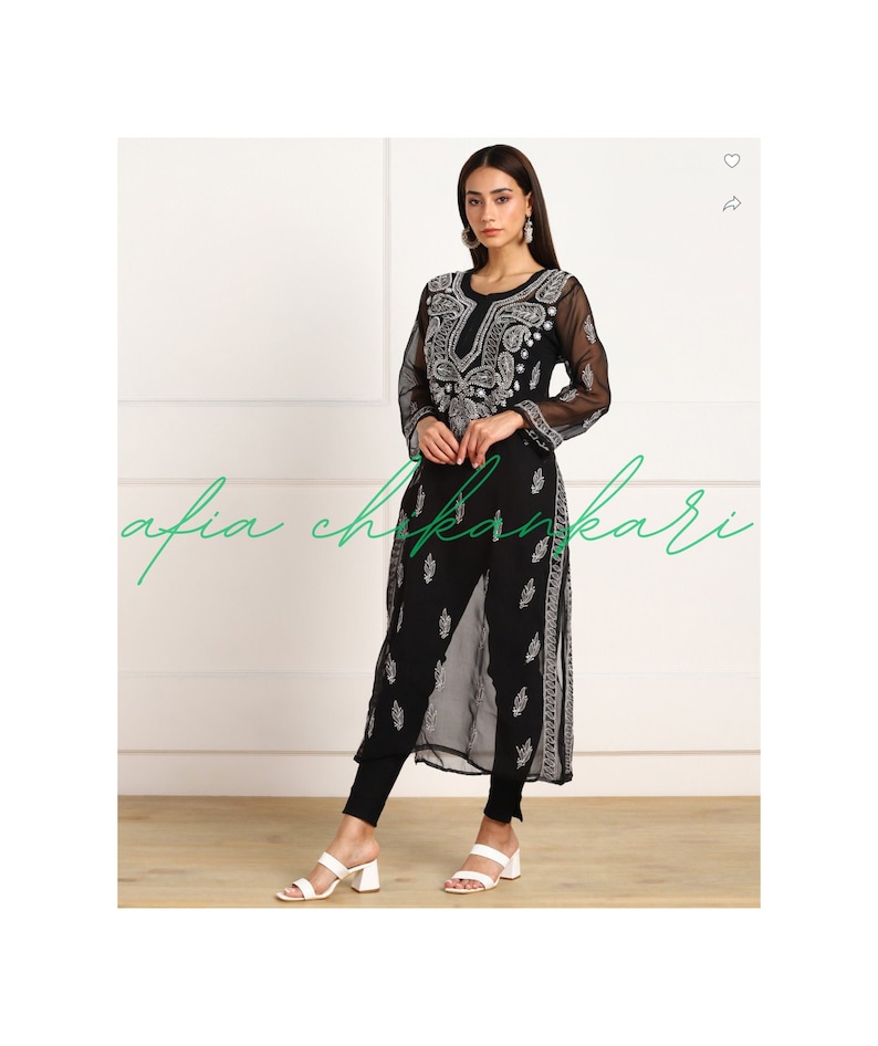 May include: A black sheer long-sleeved tunic with white embroidery. The tunic has a round neckline and a long, flowing silhouette. The model is wearing the tunic with black leggings and white sandals.