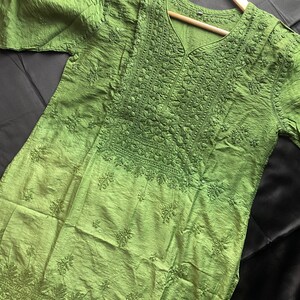 May include: A green tunic with intricate embroidery and a V-neck design. The fabric has a textured appearance, and the embroidery is concentrated around the neckline and lower hem. The tunic is displayed on a wooden hanger against a dark background.