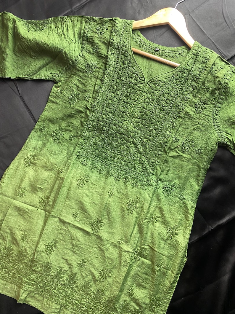 May include: A green tunic with intricate embroidery and a V-neck design. The fabric has a textured appearance, and the embroidery is concentrated around the neckline and lower hem. The tunic is displayed on a wooden hanger against a dark background.