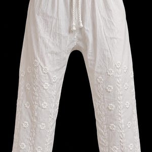 May include: White trousers featuring an elasticated waistband with a drawstring closure. The legs are decorated with vertical rows of white floral embroidery and leaf patterns, finished with a detailed textured border at the hemline.