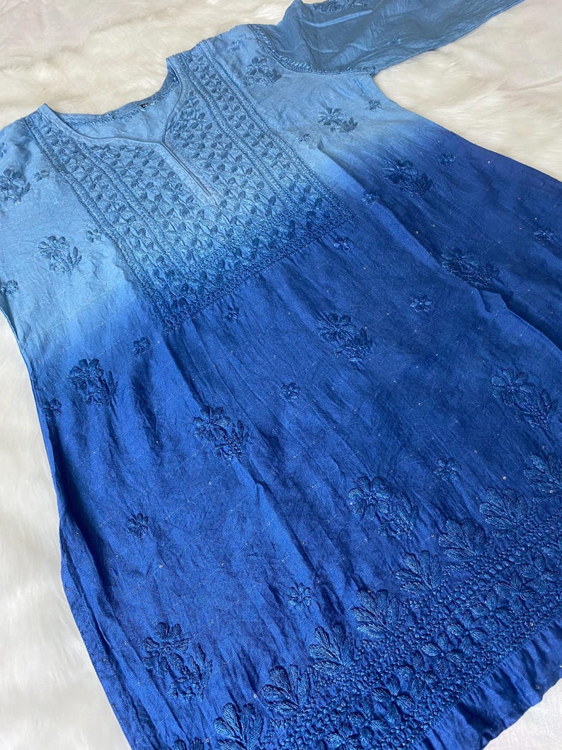 May include: A blue ombre dress with intricate floral embroidery. The dress transitions from a light blue at the top to a deep blue at the bottom. The embroidery is concentrated around the neckline, sleeves, and hem, creating a detailed and elegant look.
