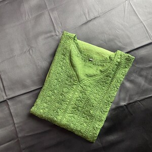 May include: A folded green tunic with a V-neck and intricate floral embroidery. The fabric has a textured appearance, and the garment is displayed on a dark, possibly black, background.