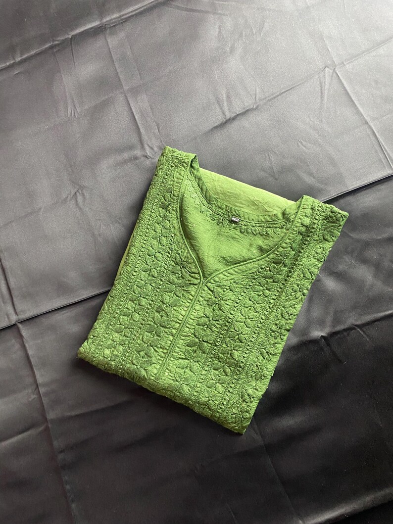 May include: A folded green tunic with a V-neck and intricate floral embroidery. The fabric has a textured appearance, and the garment is displayed on a dark, possibly black, background.