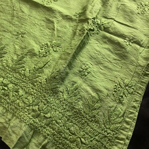 May include: Close-up of a lime green fabric with intricate embroidered floral and leaf patterns. The fabric has a textured appearance with detailed stitching along the border and scattered across the surface. The textile is likely used for clothing or home decor.