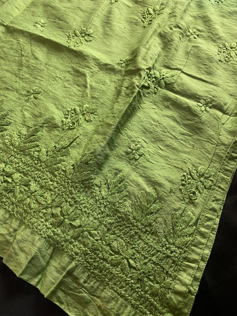 May include: Close-up of a lime green fabric with intricate embroidered floral and leaf patterns. The fabric has a textured appearance with detailed stitching along the border and scattered across the surface. The textile is likely used for clothing or home decor.