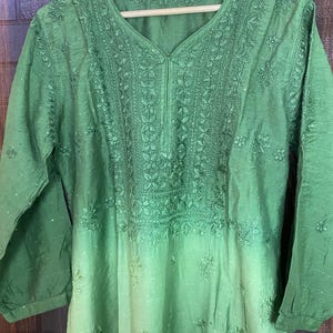 May include: A green tunic with intricate floral embroidery around the neckline and bodice. The long-sleeved top has a V-neck and a subtle ombre effect, fading from a darker green at the top to a lighter shade at the bottom. The garment is hanging on a wooden hanger.