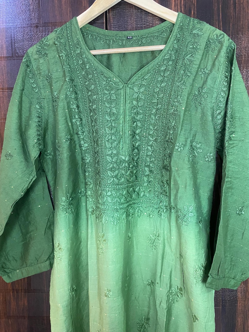 May include: A green tunic with intricate floral embroidery around the neckline and bodice. The long-sleeved top has a V-neck and a subtle ombre effect, fading from a darker green at the top to a lighter shade at the bottom. The garment is hanging on a wooden hanger.