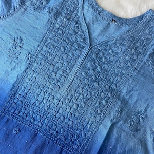 May include: A light blue tunic with intricate floral embroidery. The fabric transitions from a lighter shade at the top to a deeper blue at the bottom. The V-neckline is detailed with a vertical seam.