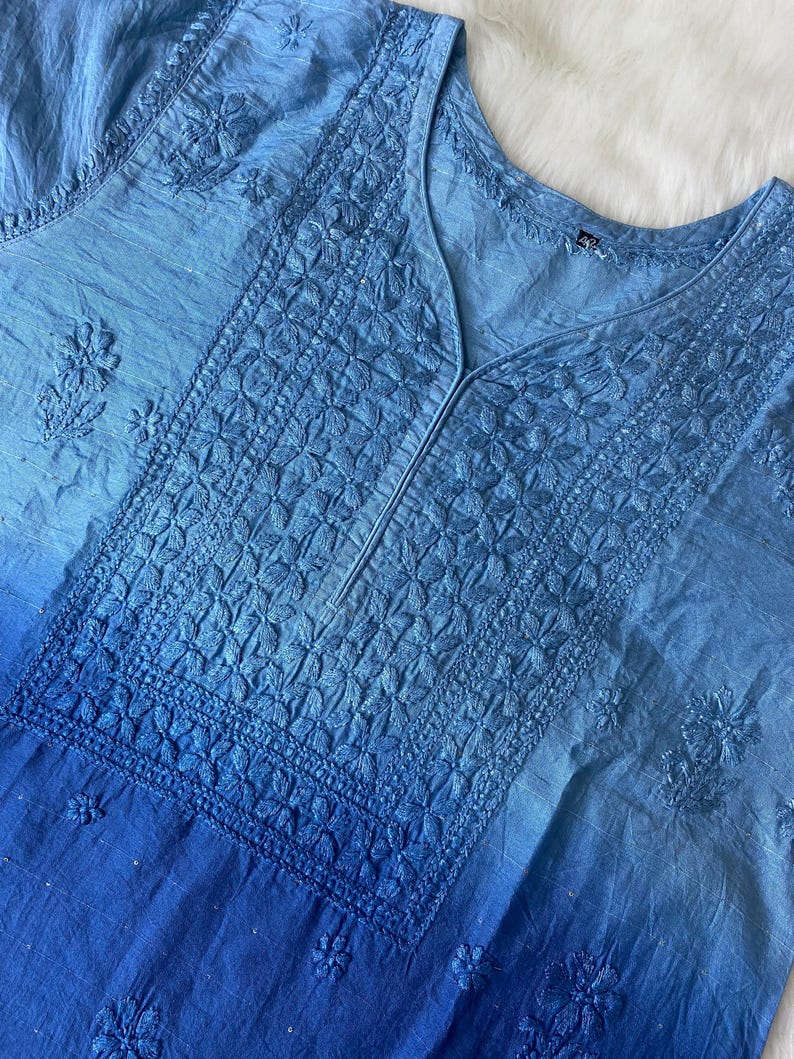 May include: A light blue tunic with intricate floral embroidery. The fabric transitions from a lighter shade at the top to a deeper blue at the bottom. The V-neckline is detailed with a vertical seam.