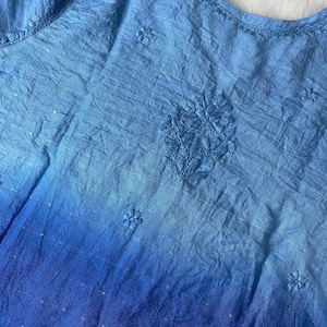 May include: A blue, sleeveless top with an ombre effect, transitioning from light to deep blue. The fabric has a crinkled texture and features embroidered floral details and delicate sequin accents. The neckline is also embroidered.