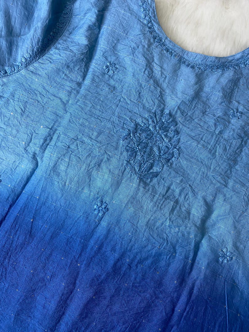 May include: A blue, sleeveless top with an ombre effect, transitioning from light to deep blue. The fabric has a crinkled texture and features embroidered floral details and delicate sequin accents. The neckline is also embroidered.