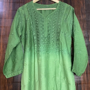 May include: A green tunic with long sleeves and intricate embroidery around the neckline and upper chest. The fabric transitions from a darker green at the top to a lighter shade towards the bottom. The tunic is hanging on a wooden hanger.