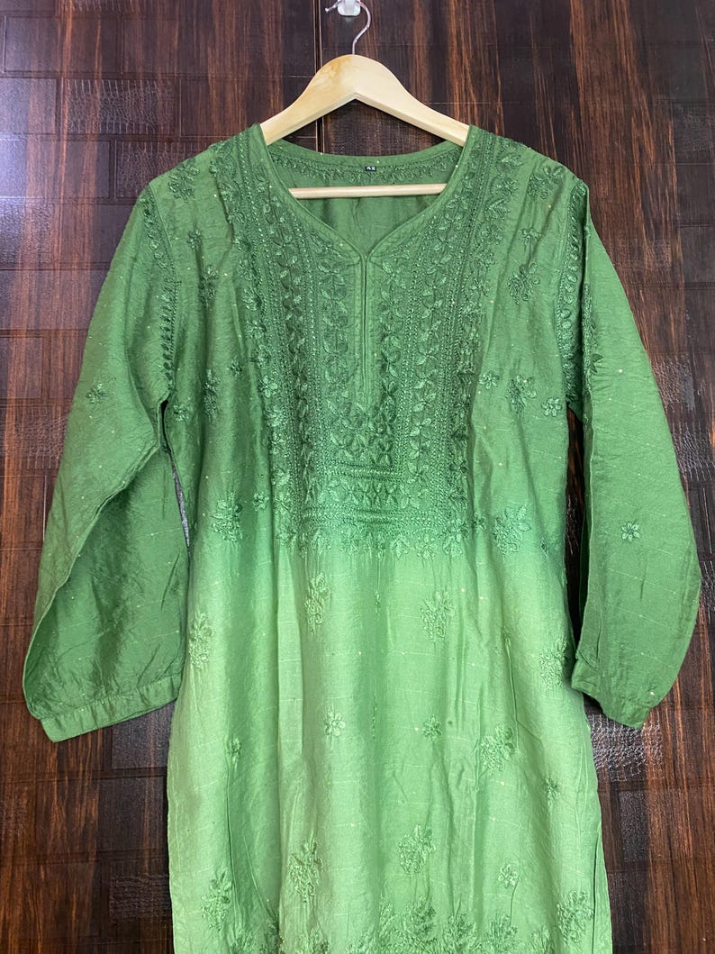 May include: A green tunic with long sleeves and intricate embroidery around the neckline and upper chest. The fabric transitions from a darker green at the top to a lighter shade towards the bottom. The tunic is hanging on a wooden hanger.