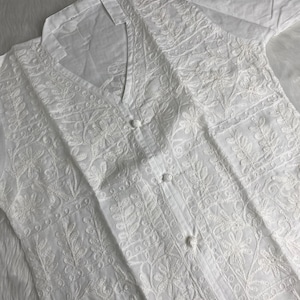 Pode incluir: Camisa branca de manga comprida com bordados florais intrincados. A camisa apresenta decote em V e fechos de botões na frente. O bordado detalhado cobre a frente e as mangas, criando um visual texturizado e elegante.