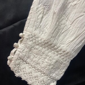 May include: Close-up of a white, long-sleeved garment with intricate embroidery and lace detailing. The sleeve cuff features a delicate crochet trim and small, decorative buttons. The fabric appears to be lightweight and textured.