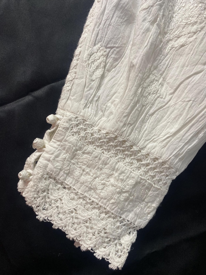May include: Close-up of a white, long-sleeved garment with intricate embroidery and lace detailing. The sleeve cuff features a delicate crochet trim and small, decorative buttons. The fabric appears to be lightweight and textured.