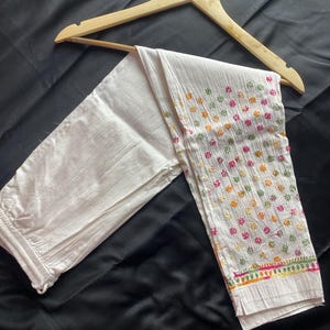 Può includere: Pantaloni bianchi ricamati con motivi floreali colorati, appesi a una gruccia di legno. I pantaloni hanno un bordo ricamato dettagliato con accenti rosa, verdi e arancioni. Il tessuto sembra leggero e adatto al clima caldo.
