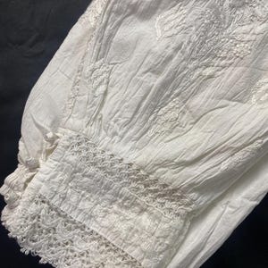May include: Close-up of white fabric with intricate floral embroidery and a delicate lace trim. The fabric appears to be slightly wrinkled, suggesting a soft, natural material. The design features detailed stitching and a textured appearance.