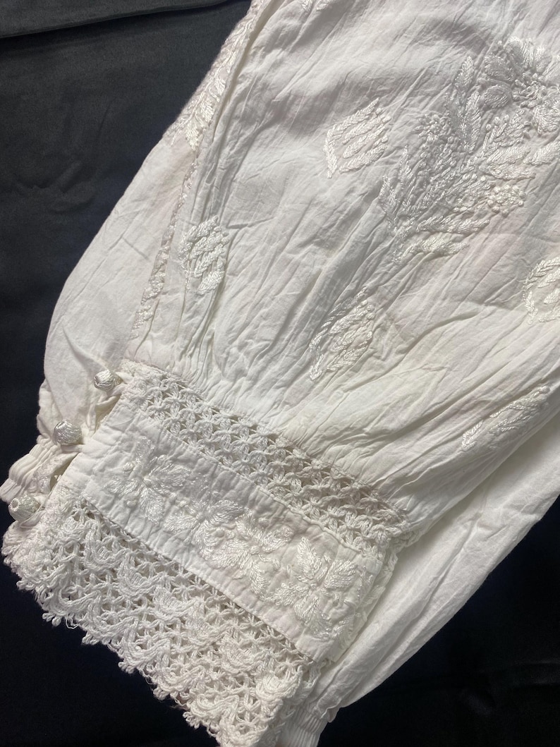 May include: Close-up of white fabric with intricate floral embroidery and a delicate lace trim. The fabric appears to be slightly wrinkled, suggesting a soft, natural material. The design features detailed stitching and a textured appearance.