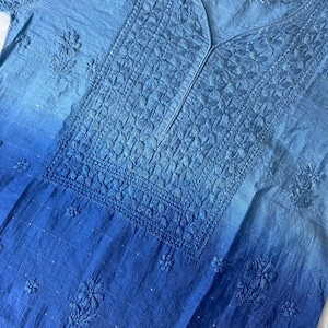 May include: A light blue tunic with intricate floral embroidery. The fabric transitions from a lighter shade at the top to a deeper blue at the bottom. The neckline is a V-shape, and the sleeves are short.