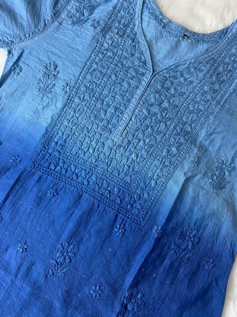 May include: A light blue tunic with intricate floral embroidery. The fabric transitions from a lighter shade at the top to a deeper blue at the bottom. The neckline is a V-shape, and the sleeves are short.
