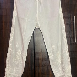 May include: White, lightweight pants with intricate embroidered floral patterns along the sides and at the cuffs. The pants have a drawstring waist and a relaxed fit, perfect for casual wear. The fabric appears to be a soft, breathable material.