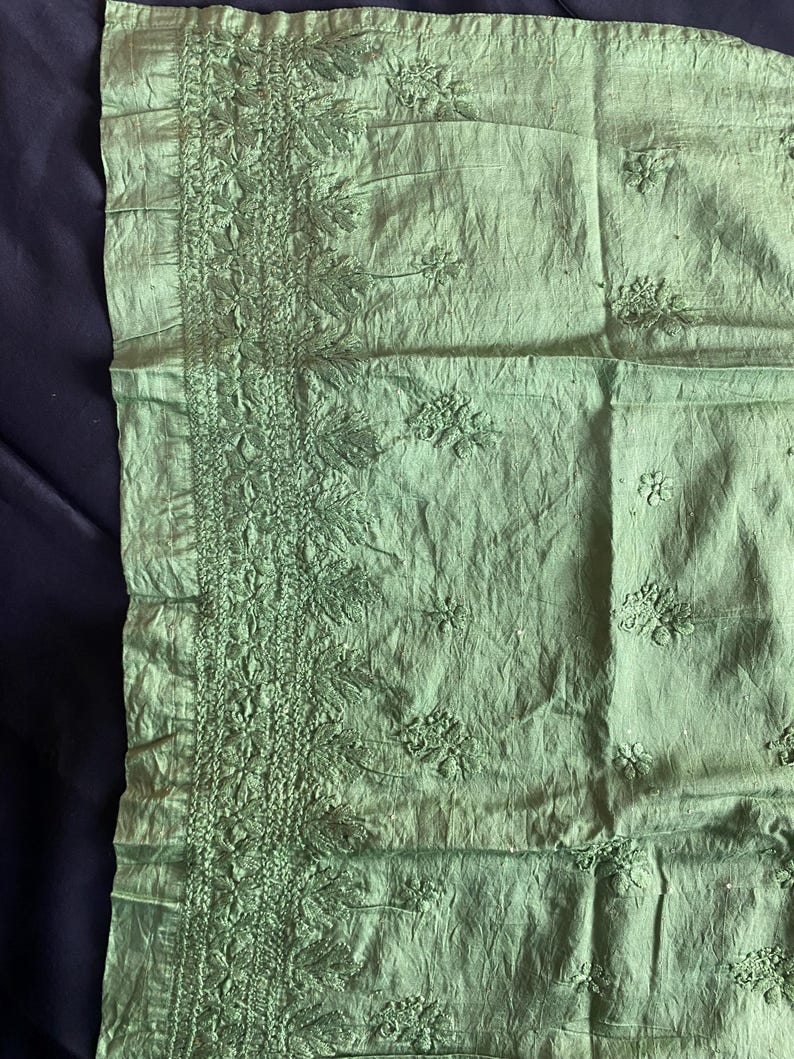 May include: A light green textile with intricate floral embroidery along one edge. The fabric has a slightly wrinkled texture, and the embroidery features detailed leaf and flower patterns. The overall aesthetic is delicate and handcrafted.
