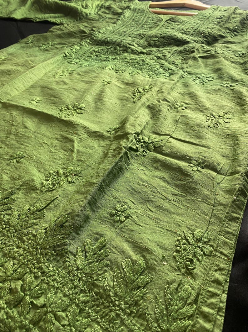 May include: A lime green tunic with intricate floral embroidery. The long-sleeved garment features detailed stitching around the neckline, sleeves, and hem. The fabric has a slightly textured appearance, and the overall design is elegant and detailed.