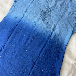 May include: A blue ombre dress with short sleeves. The dress features embroidered floral details and a textured fabric. The color transitions from light blue at the top to a deep blue at the bottom. The dress is laid on a white, fluffy surface.