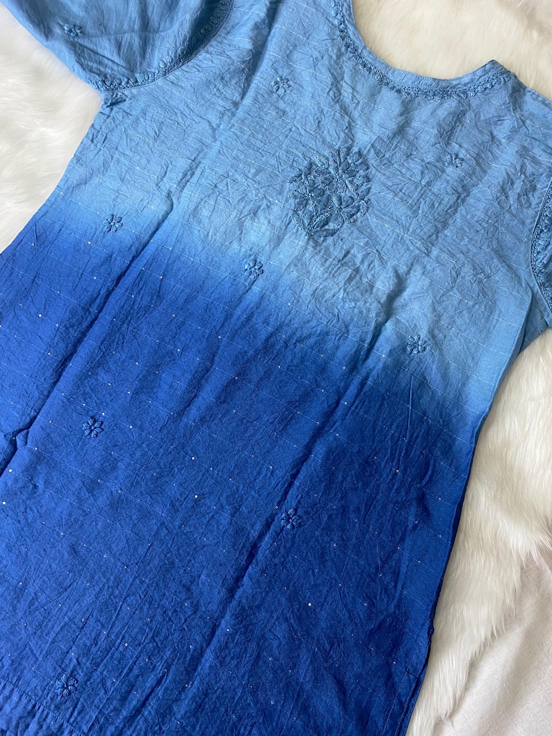 May include: A blue ombre dress with short sleeves. The dress features embroidered floral details and a textured fabric. The color transitions from light blue at the top to a deep blue at the bottom. The dress is laid on a white, fluffy surface.