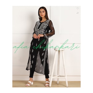 May include: A black sheer long-sleeved tunic with white embroidery. The tunic has a round neckline and a straight hem. The model is wearing black trousers and white sandals.