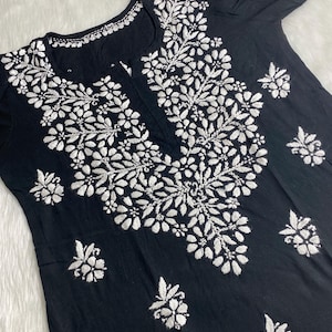以下が含まれることがあります： 白い刺繍が施された黒い綿のチュニック。刺繍はネックラインを中心に、チュニックの前身頃まで伸びています。