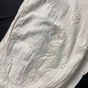 May include: Close-up of a white, textured fabric with embroidered floral and leaf patterns. The fabric has a crinkled appearance and a crocheted trim at the bottom. The background is dark.