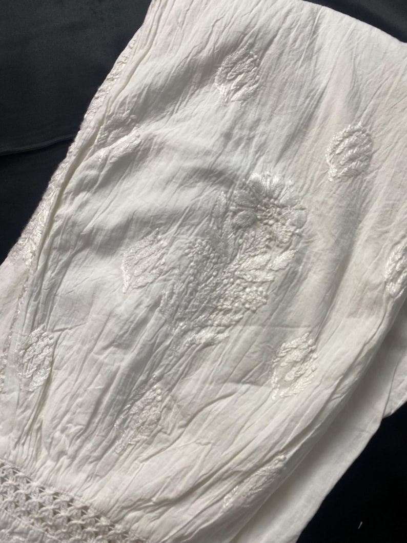 May include: Close-up of a white, textured fabric with embroidered floral and leaf patterns. The fabric has a crinkled appearance and a crocheted trim at the bottom. The background is dark.