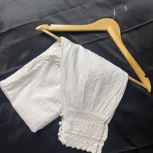 May include: White, textured fabric pants with a decorative lace trim at the ankle. The pants are displayed on a wooden hanger against a black background. The fabric appears to be lightweight and suitable for warm weather.